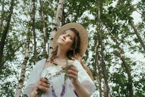 A woman in a straw hat enjoys a sunny day in a lush forest, holding wildflowers.